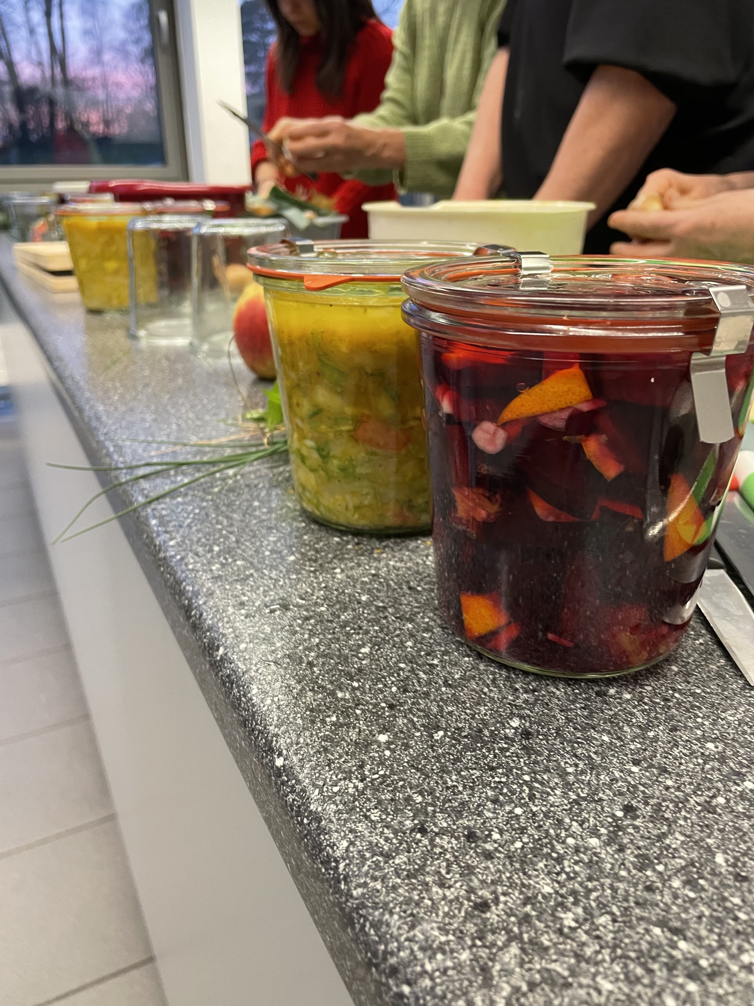 Mehrere eingelegte Gemüsegläser auf Küchentheke, Personen schneiden im Hintergrund, Fischl Biohof Niederbayern